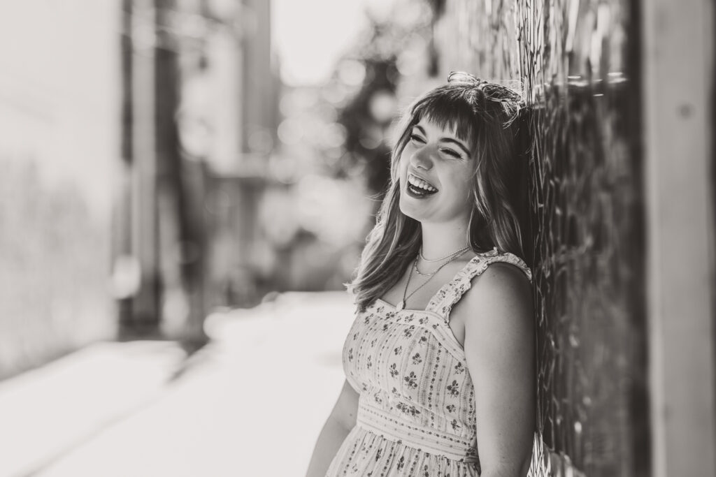 Black and white candid senior portrait of Lyla laughing while leaning against a textured wall.