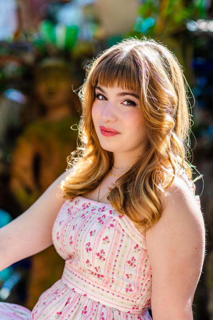 Senior portrait of Lyla looking at the camera with a soft expression against a colorful background. The sunlight is lighting the top of her red hair.