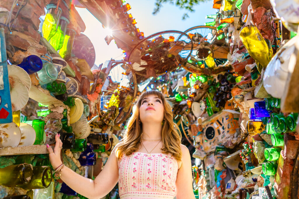 Senior portrait of a student looking upward while standing between mosaic walls decorated with bottles, tiles, and hanging art. There is a sunburst lighting the top of the mosaic art.