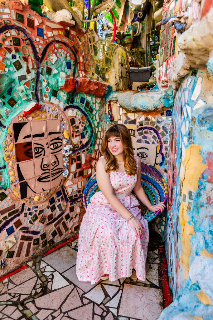 High school senior smiling while seated on a woven chair, surrounded by vibrant mosaic artwork and tile faces.
