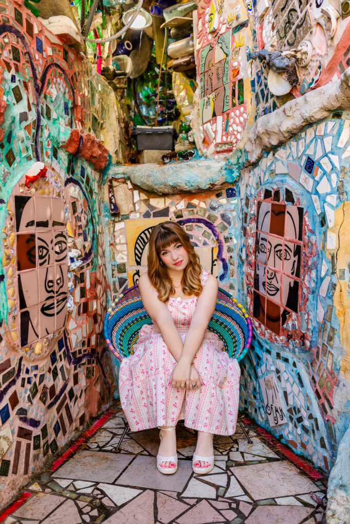 Layla, a high school senior with red hair with bangs wearing a white dress with pink flowers, is sitting on a colorful woven chair surrounded by mosaic artwork at Philadelphia’s Magic Gardens. Her hands are together over her knees as she softly stares at the camera and with her head tilted.