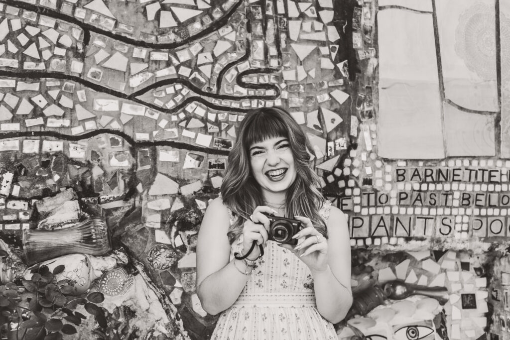 Black and white senior portrait of Lyla smiling while holding her camera in front of a mosaic wall.
