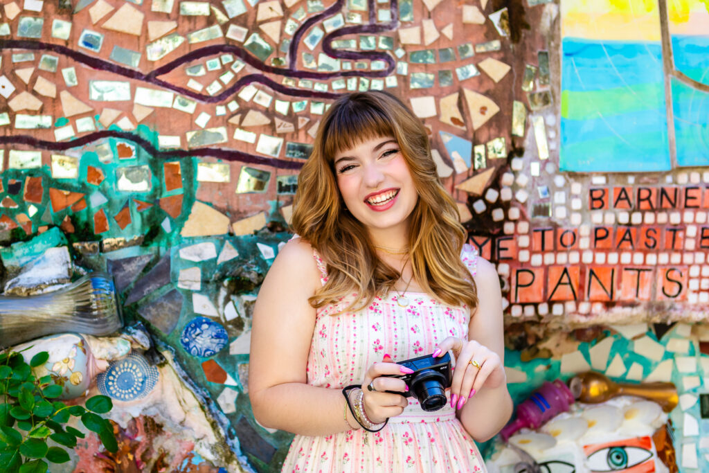 Portrait of Lyla smiling while holding her camera against a colorful mosaic wall during her Philadelphia Senior Session.