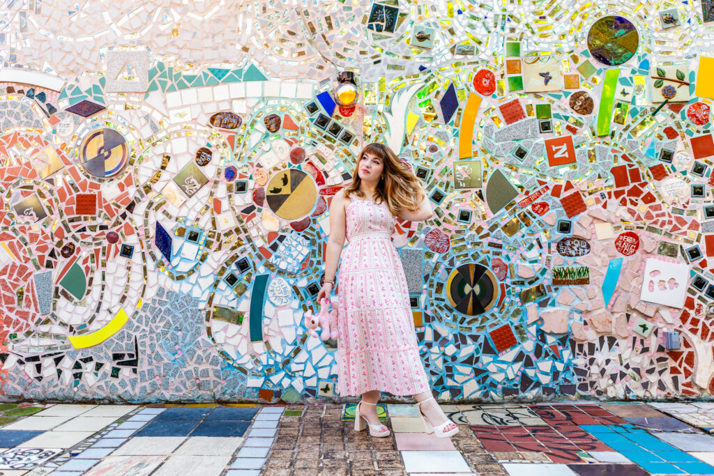 Full-body senior portrait of Lyla standing in front of a colorful mosaic wall at the Magic Gardens. Lyla is kicking one leg out while wearing a white sundress with pink flowers and holding a childhood stuffed animal in one hand while the other hand plays with her hair.