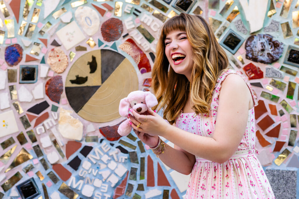 Senior portrait of Lyla laughing while holding her childhood stuffed animal against a mosaic backdrop at The Magic Gardens in Philadelphia.