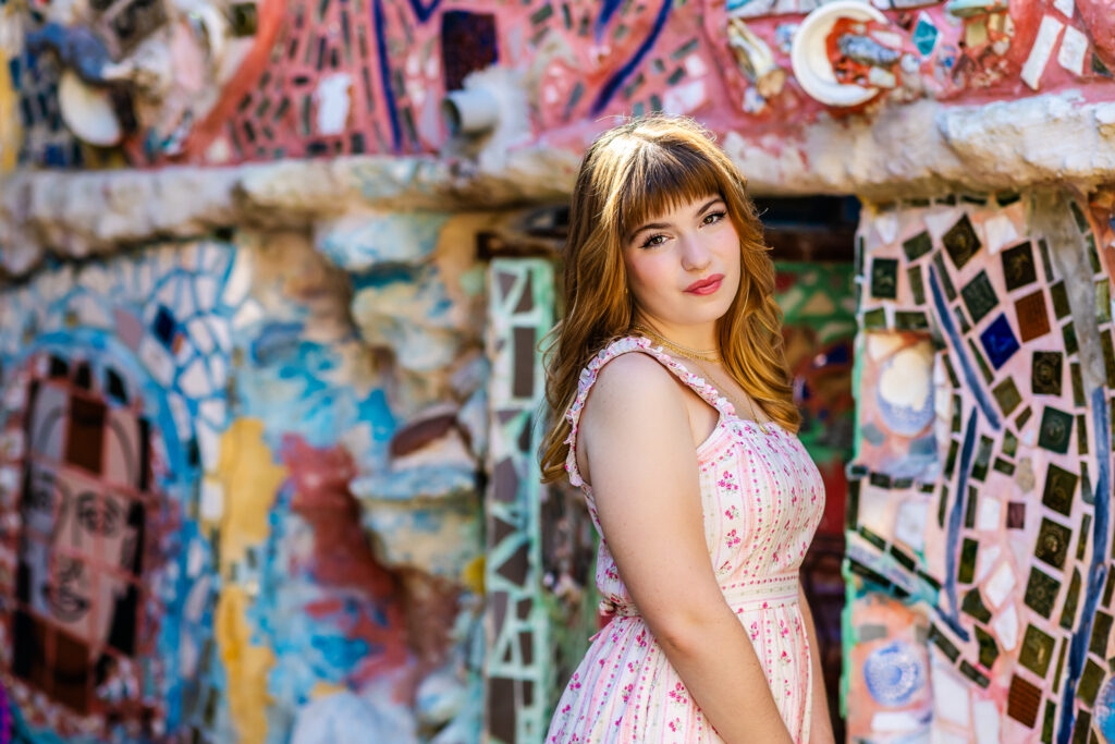Side profile portrait of a high school student with red hair and bangs and a pink and white dress standing among textured mosaic walls and colorful tiles during a Philadelphia senior session.