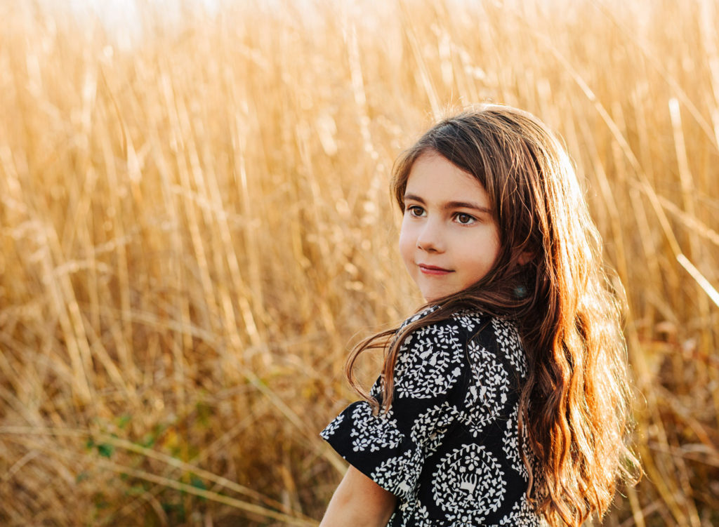 Young girl in black dress in a field of tall grass at sunset, looking off into the distance.