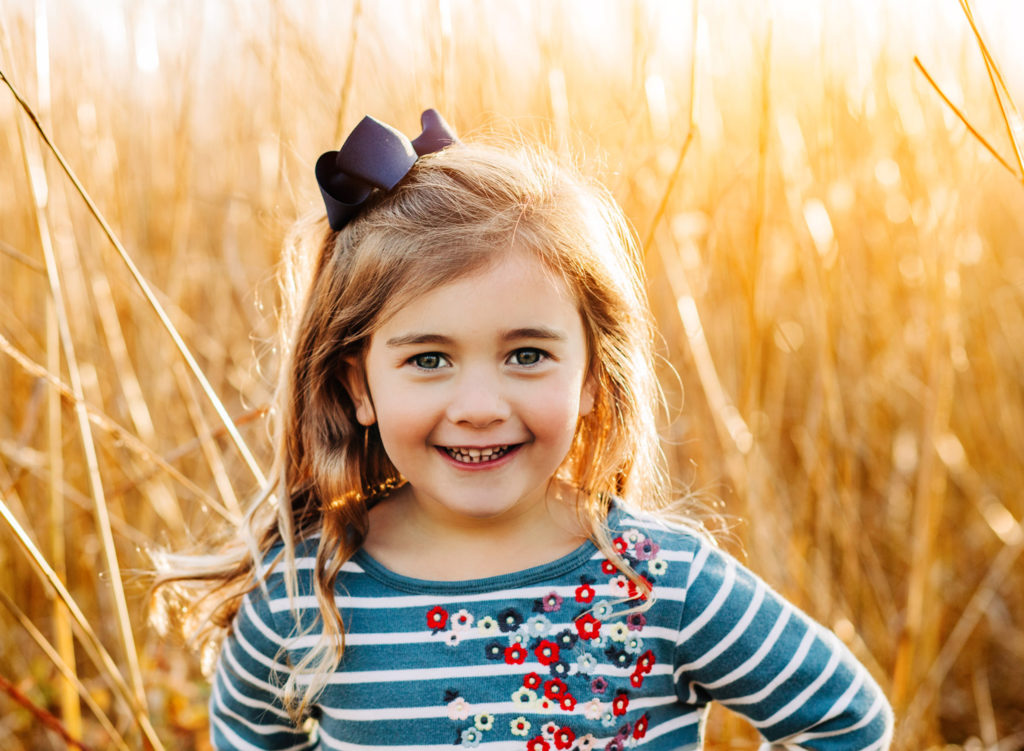 Young girl in blue striped dress in a field of tall grass at sunset, looking at the camera and smiling.