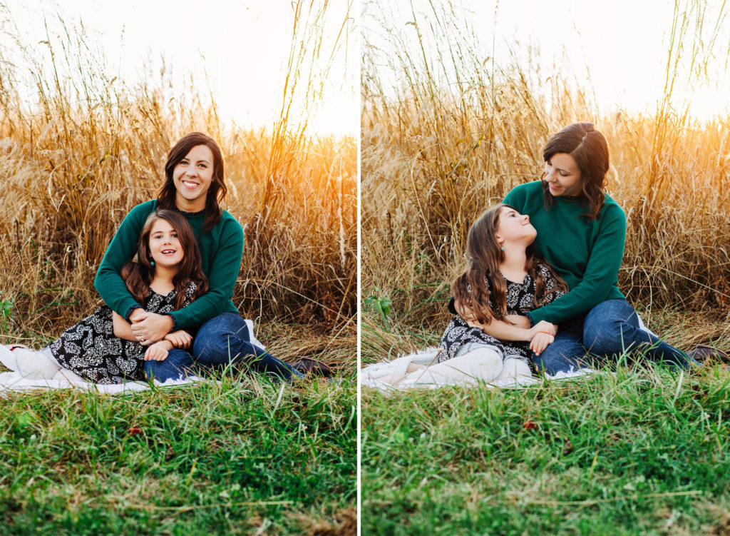 A mother and her young daughter sitting on a blanket in a field of tall grass at sunset. 