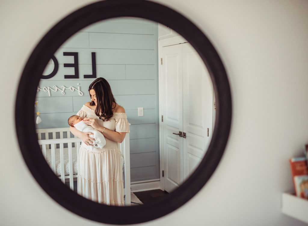 A reflection in a mirror of a  mother in a long white dress holding a swaddled newborn baby.
