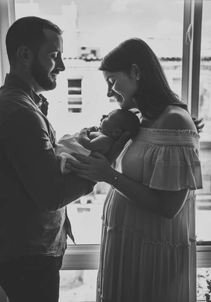 A black and white image of a father and mother holding their newborn baby swaddled son in front of a big window.