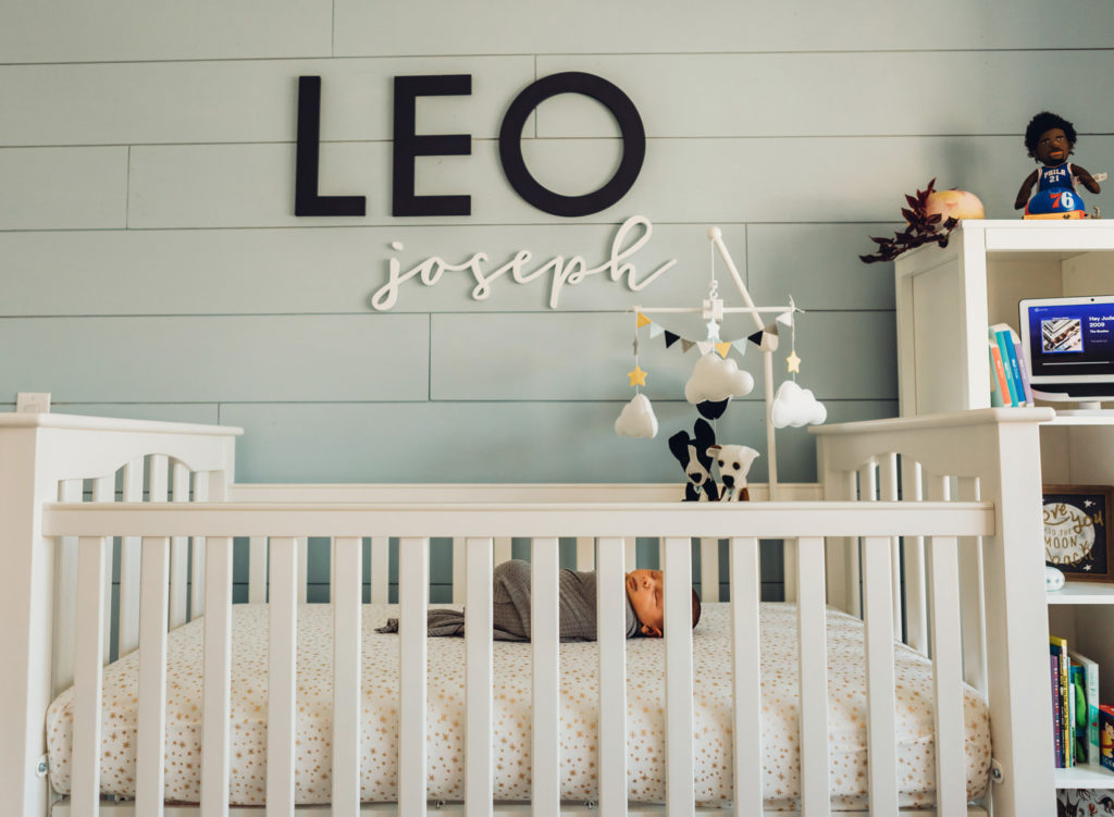 A newborn baby wrapped in a grey swaddle sleeping in a crib in a nursery. Above the bed is his name on the wall: Leo Joseph.