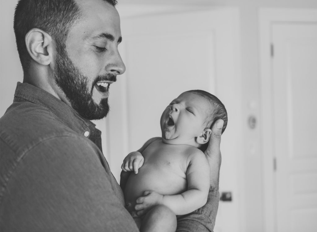A black and white image of a father holding a newborn baby up while the baby yawns.