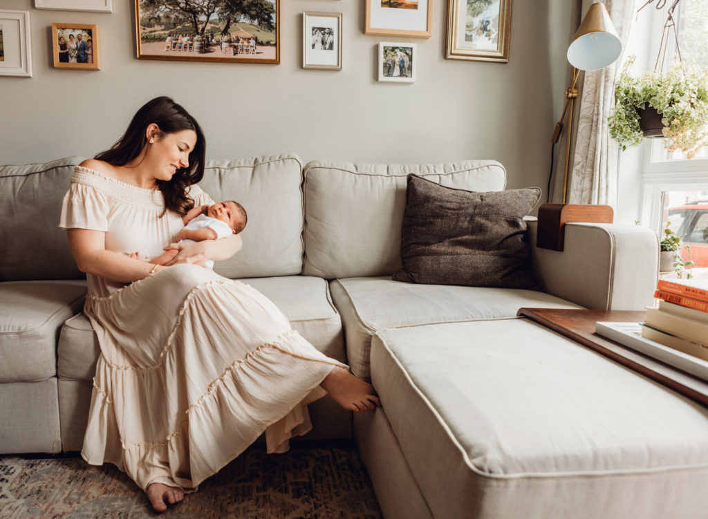 A mother holding her newborn son on her beige couch, in her living room. She is in a long beige dress and there are many framed pictures above her head on the wall.