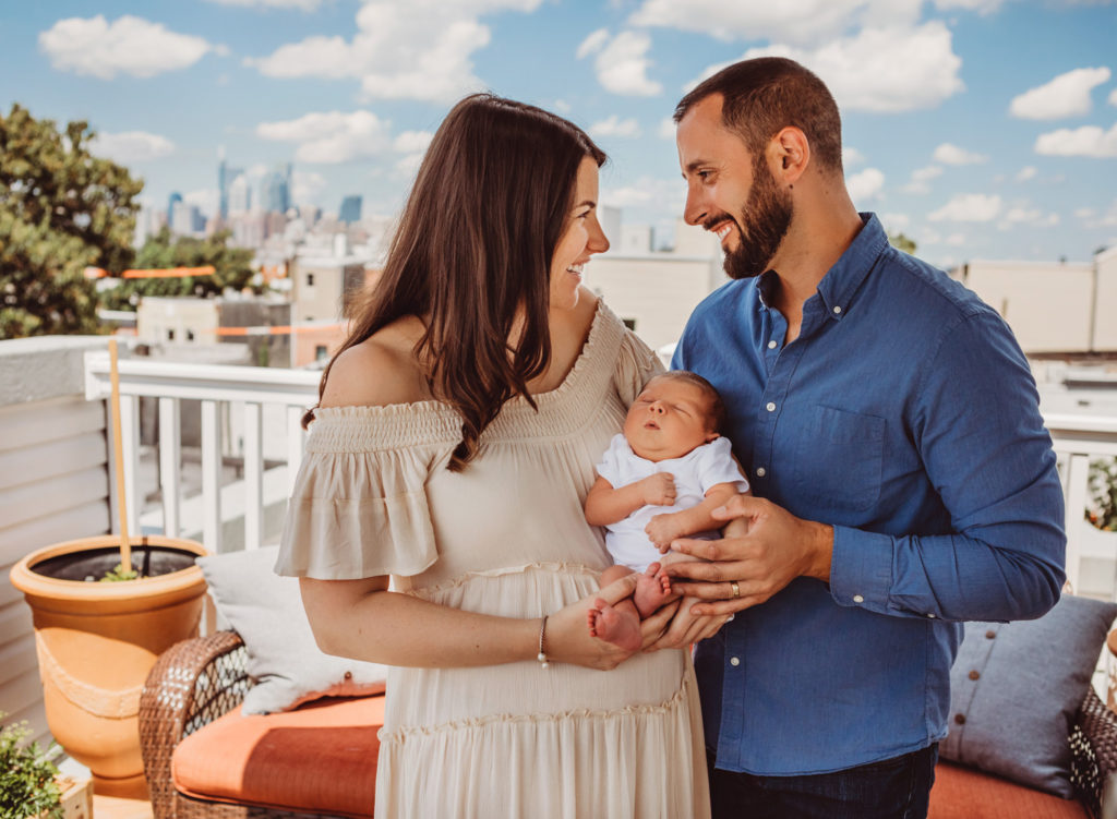 A mother and father on a rooftop deck holding their newborn baby and smiling at each other. The city of Philadelphia is in the background.
