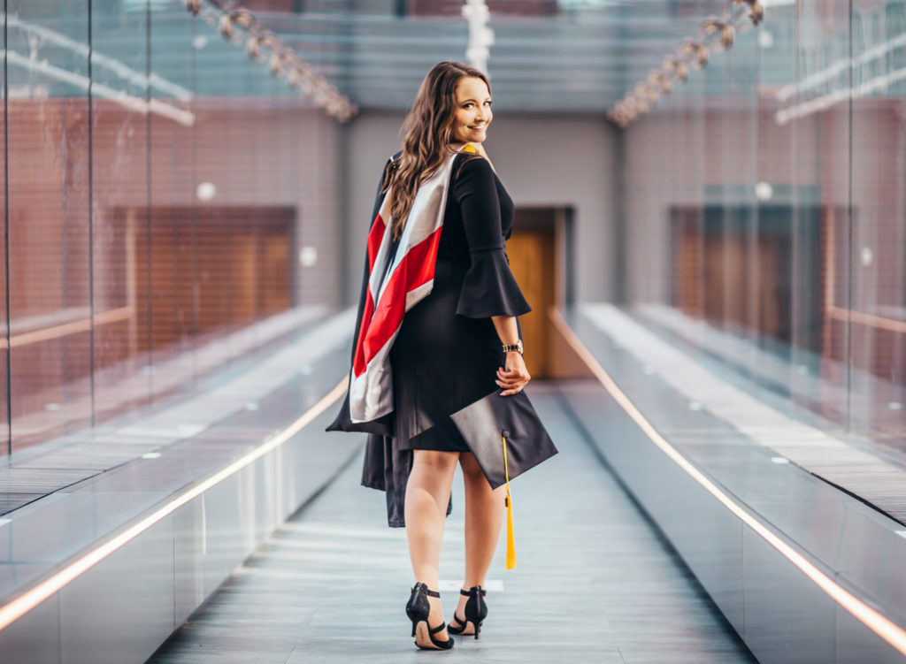 A young woman walks down a hallway of glass windows while holding her graduation cap in her hand and looking over her shoulder.