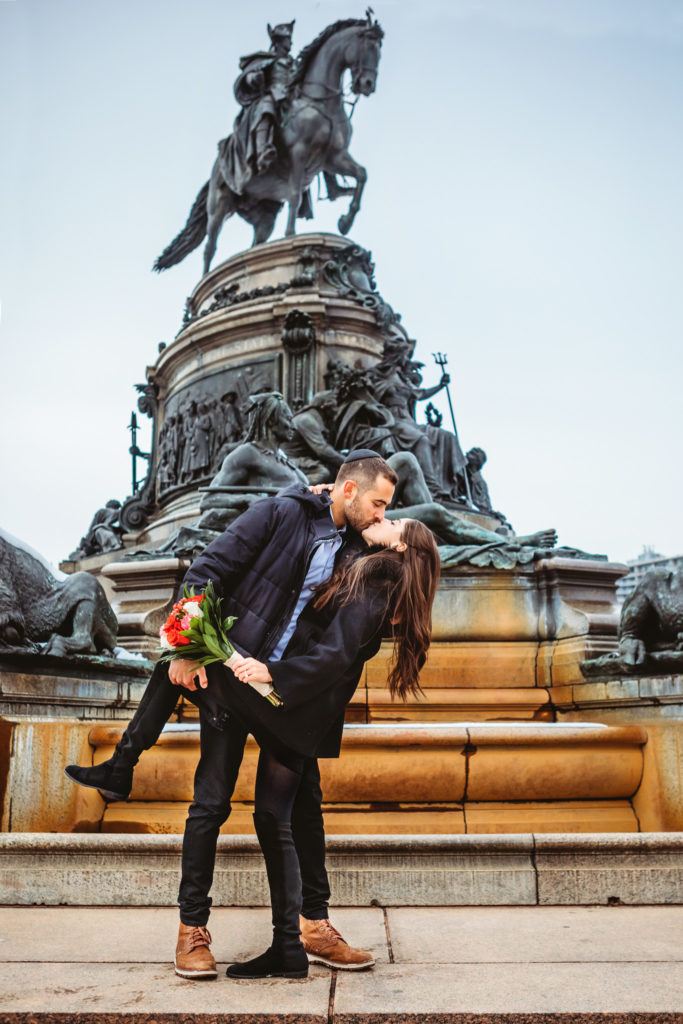 A man dipping and kissing his new fiancé in front of the Washington Monument Statue in Philadelphia on a winter day. 