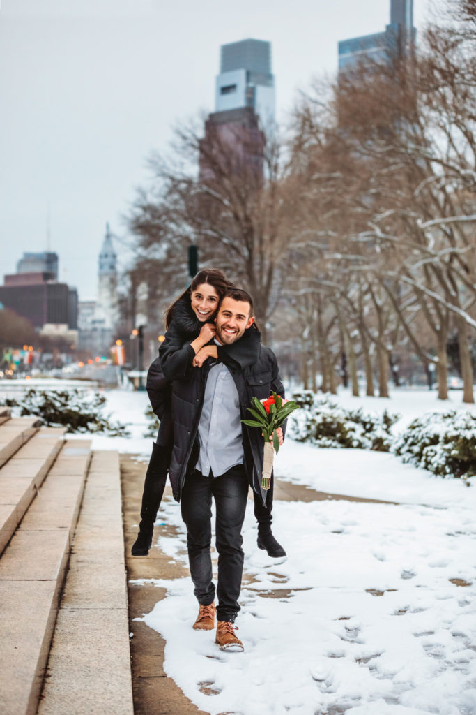 A man giving his new fiancé a piggyback ride through the snow. The city of Philadelphia is in the background. 
