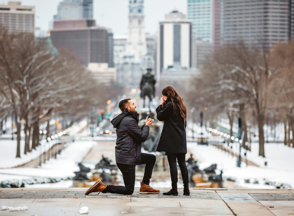 A man gets on his knee to propose to his girlfriend on the top of the steps of the front of the Philadelphia Museum of Art on a winter day. This girlfriend is covering her face while she cries.
