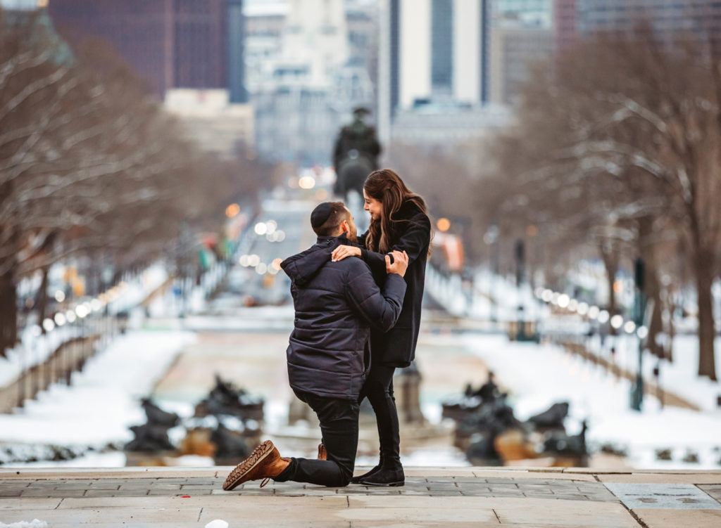 A man gets on his knee to propose to his girlfriend on the top of the steps of the front of the Philadelphia Museum of Art on a winter day. The girlfriend is crying.