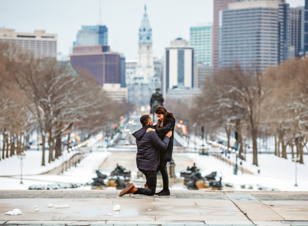 A man gets on his knee to propose to his girlfriend on the top of the steps of the front of the Philadelphia Museum of Art on a winter day. The girlfriend is crying. The city of Philadelphia and City Hall is behind them in the distance.
