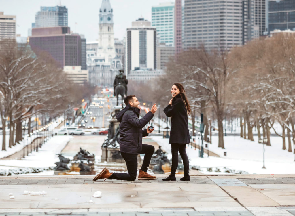 A man gets on his knee to propose to his girlfriend on the top of the steps of the front of the Philadelphia Museum of Art on a winter day. The girlfriend is laughing as the man is pointing back to the photographer. The city of Philadelphia and City Hall is behind them in the distance.
