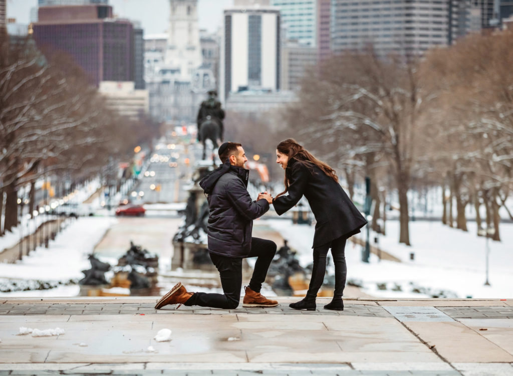 A man gets on his knee to propose to his girlfriend on the top of the steps of the front of the Philadelphia Museum of Art on a winter day. The girlfriend is holding his hands and laughing. The city of Philadelphia is behind them in the distance.