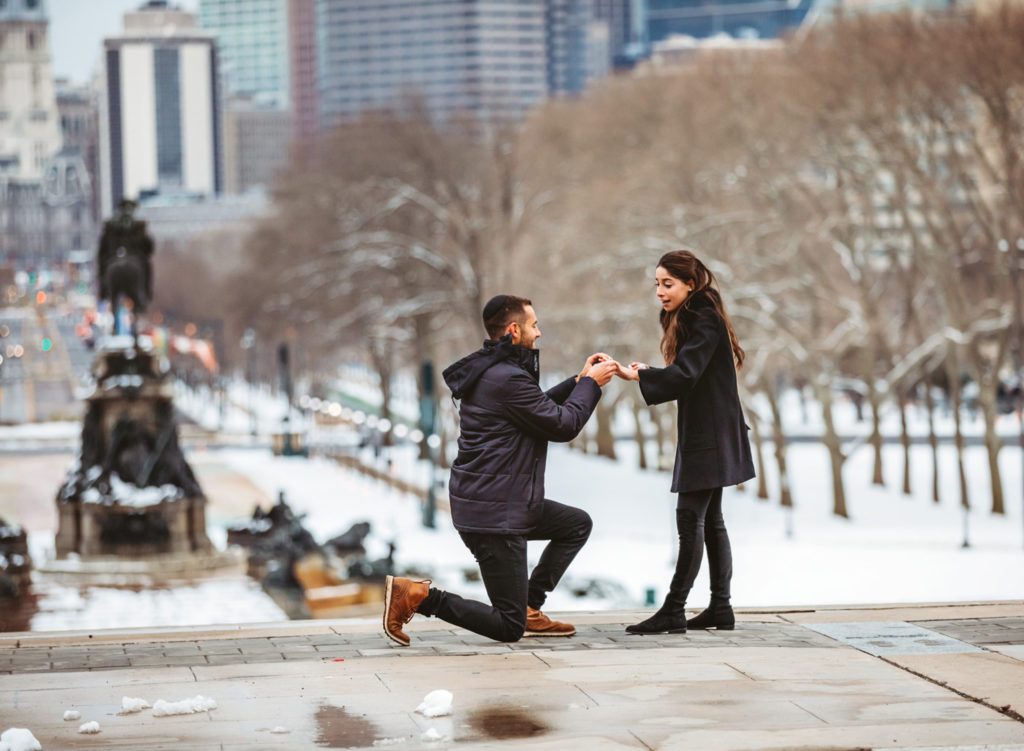 A man is on his knees proposing to his girlfriend on the top of the stairs of the Philadelphia Museum of Art. He is putting the ring on his girlfriend's finger with the city of Philadelphia in the background.
