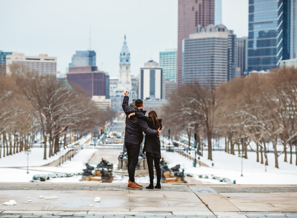 A man and his girlfriend are looking at the city of Philadelphia from the top of the steps of the Philadelphia Museum of Art on a winter day. The man has his hand triumphantly in the air after a proposal. There is snow on the ground. 