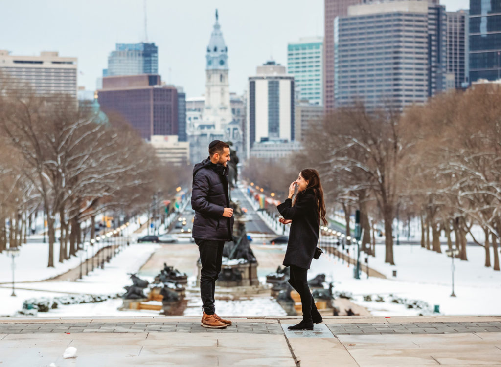 A man and woman laugh after a proposal at the top of the stairs at the Philadelphia Museum of Art. It is a winter day and the city of Philadelphia and City Hall is behind them in the distance. There is snow on the ground.