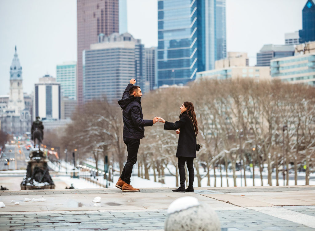 A man and his girlfriend are looking at the city of Philadelphia from the top of the steps of the Philadelphia Museum of Art on a winter day. The man has his hand triumphantly in the air after a proposal. There is snow on the ground. 