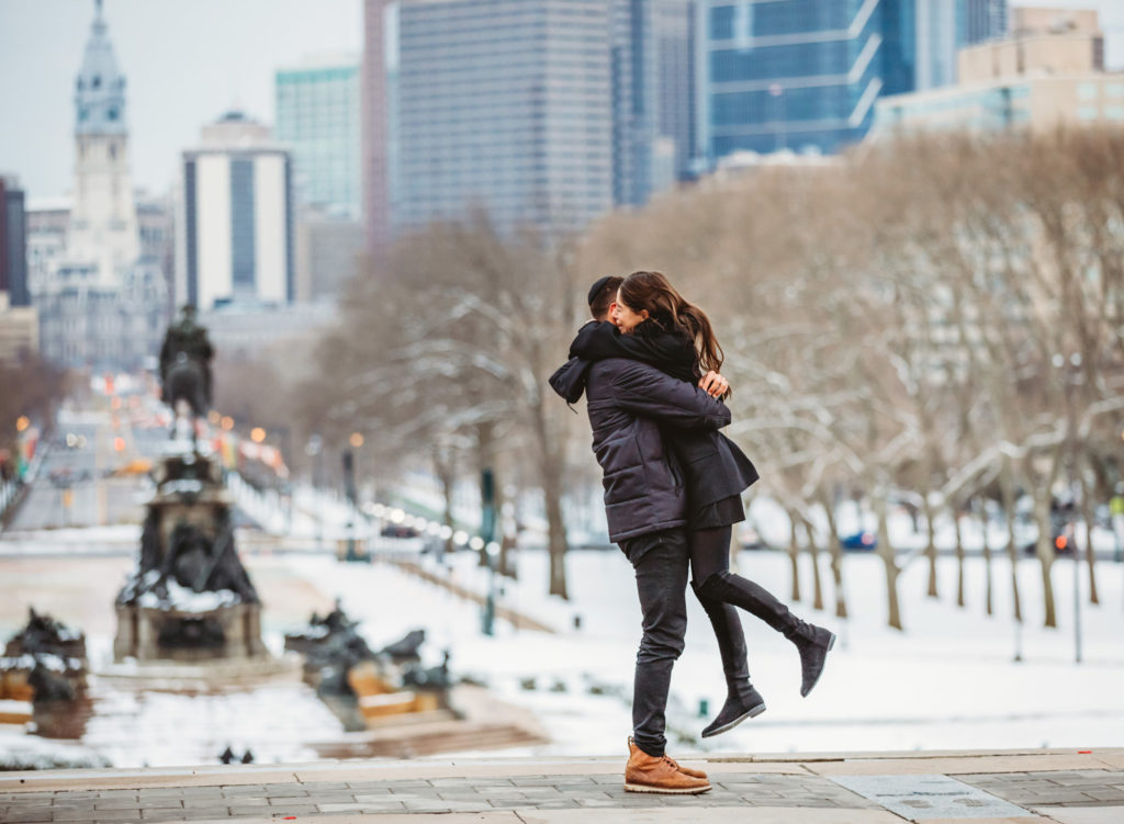 A man and woman hug after a proposal on top of the steps in front of the Philadelphia Museum of Art on a winter day.