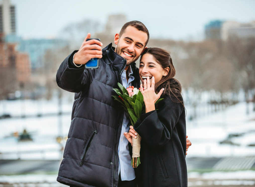 A man and woman take a selfie while the woman shows off her engagement ring after a surprise proposal at the top of the stairs at the Philadelphia Museum of Art. They are both smiling and laughing. 