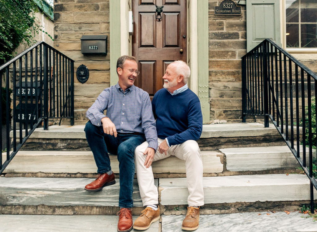 Two men, dressed in blue casual clothes, sit closely on front steps of a home together as they look at one another and laugh.