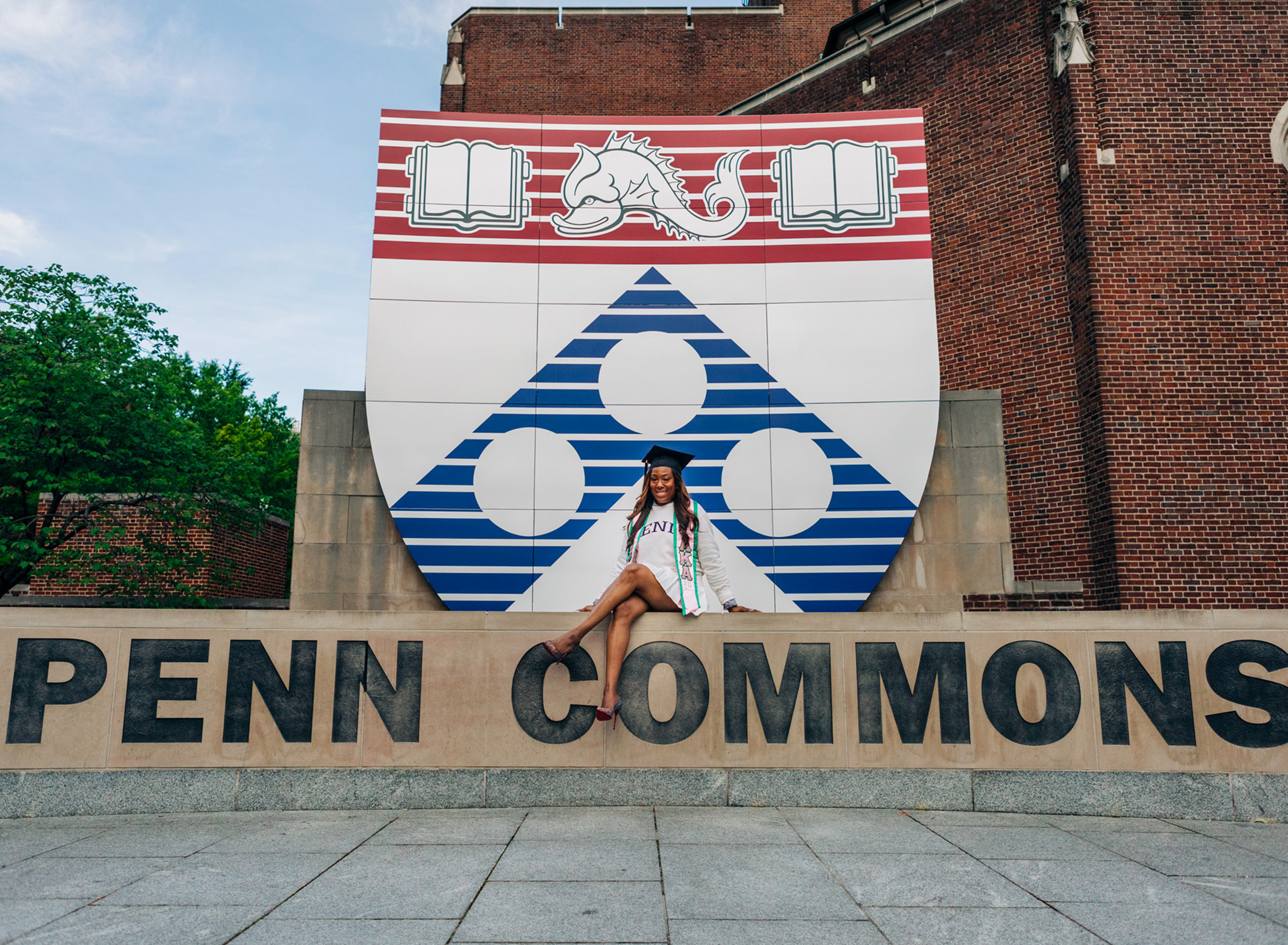 African American woman sits on a ledge at Penn Commons at the University of Pennsylvania in a white tennis outfit and a graduation cap