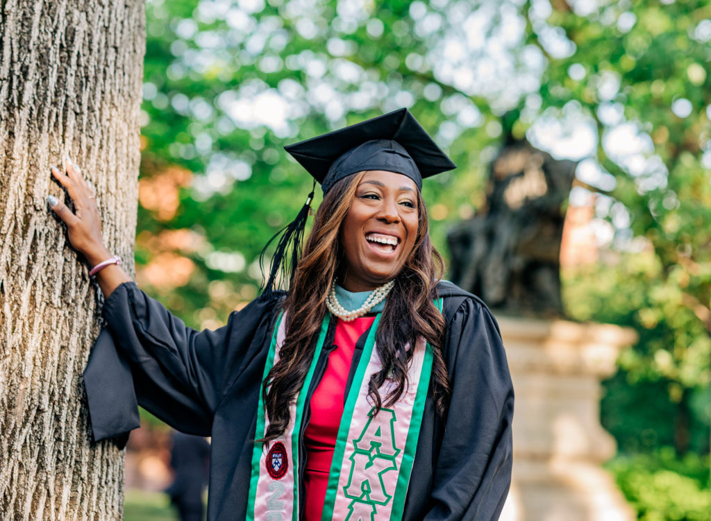 African American woman in a graduation cap and gown leans against a tree and laughs during a graduation photo session.

