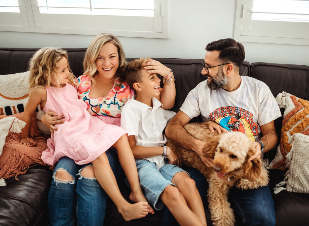 A family of four and their golden doodle do sitting together on a leather couch looking at each other and laughing. 