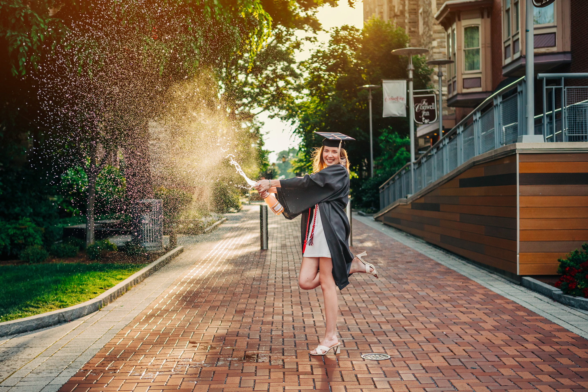 A female college graduate with long red hair in a white dress with a black cap and gown is on the Temple University campus. She is spraying a champagne bottle in celebration with the golden hour sun shining behind her.