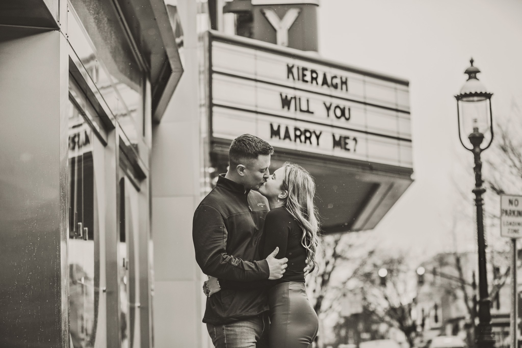 A black and white image of young man kissing a young woman under the marquee sign of the County Theater in Doylesotown, Pa in the rain. The marquee reads, "Kieragh, will you marry me?"