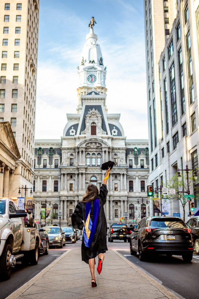 Woman walking away from the camera in a graduation robe in front of City Hall in Philadelphia as she's holding her cap in the air.