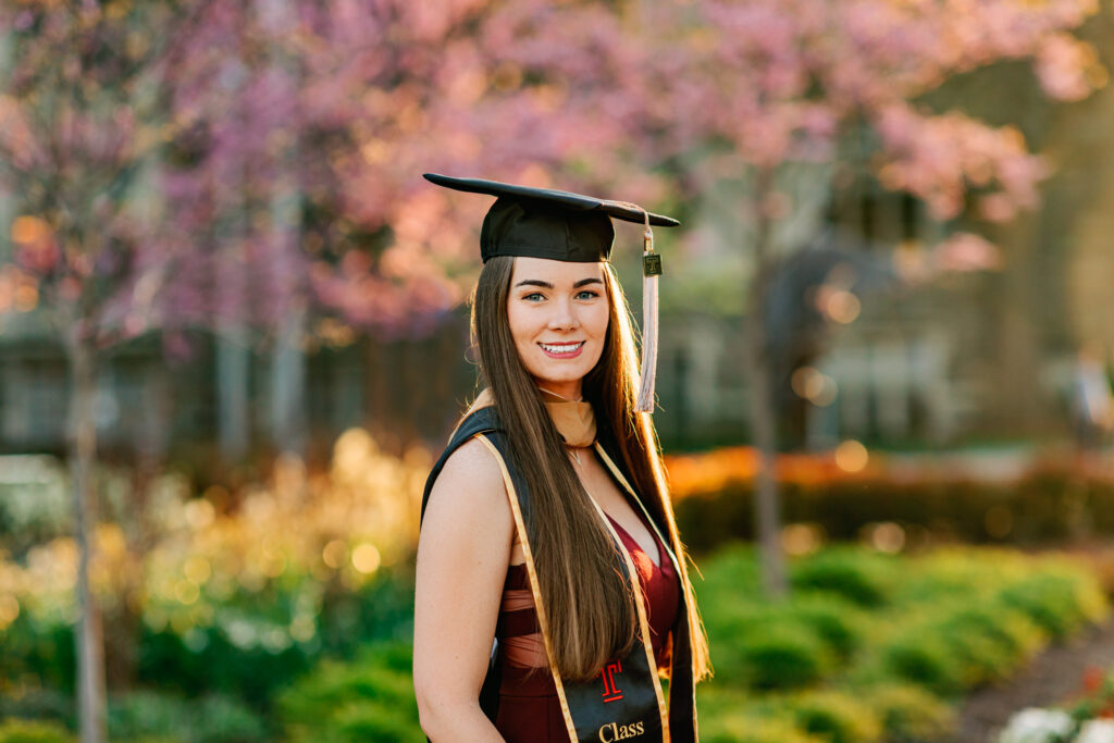 Graduate wearing a cap and stole smiles during a spring portrait session with blooming trees behind her.