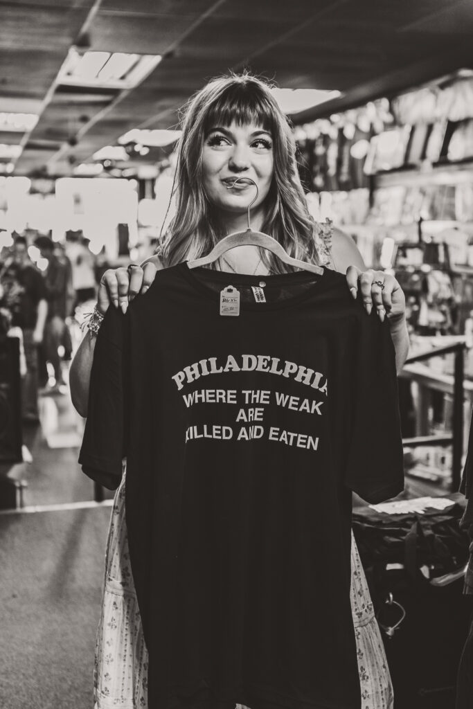 Black and white senior portrait of Lyla holding a graphic t-shirt that reads "Philadelphia, where the weak are killed and eaten" inside a record shop in Philadelphia