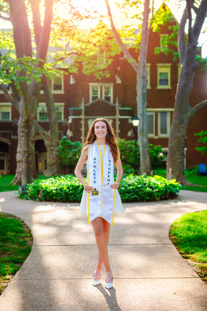 College graduate wearing athlete and student stoles standing on a tree-lined campus walkway at golden hour.