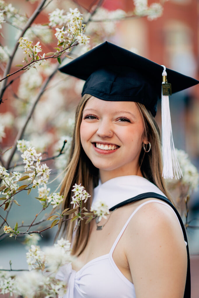 Graduate in cap and gown smiling while standing among blooming white flowers on campus.