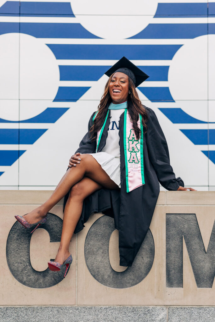 Graduate sitting with legs crossed and confidently laughing in cap and gown during a graduation photoshoot in Philadelphia.