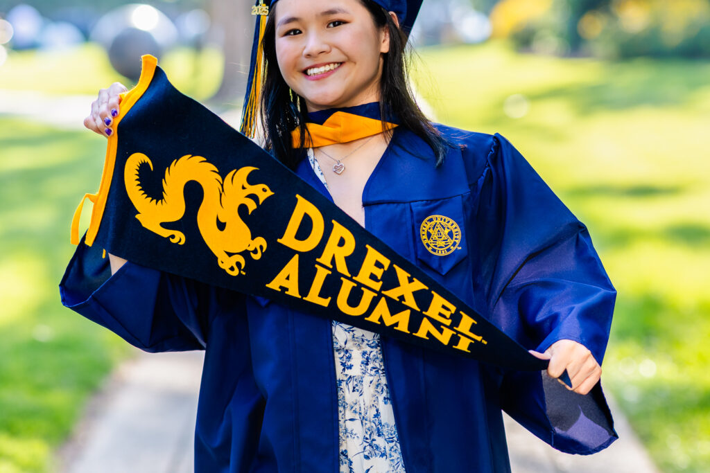 Drexel University graduate holding a Drexel Alumni banner while smiling in a sunlit outdoor setting.