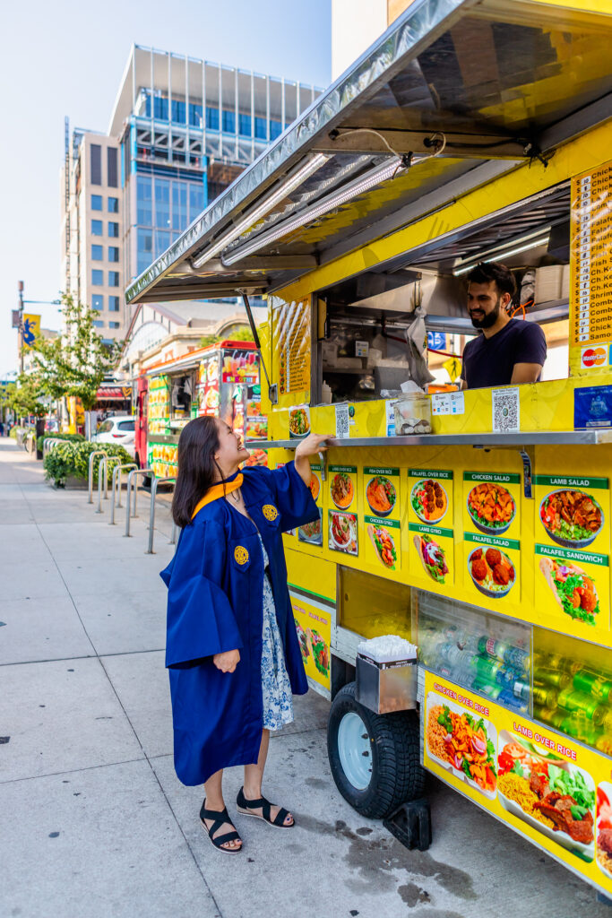 Graduate wearing a blue graduation gown ordering food at a colorful food truck on a city sidewalk, on the Drexel University campus in Philadelphia