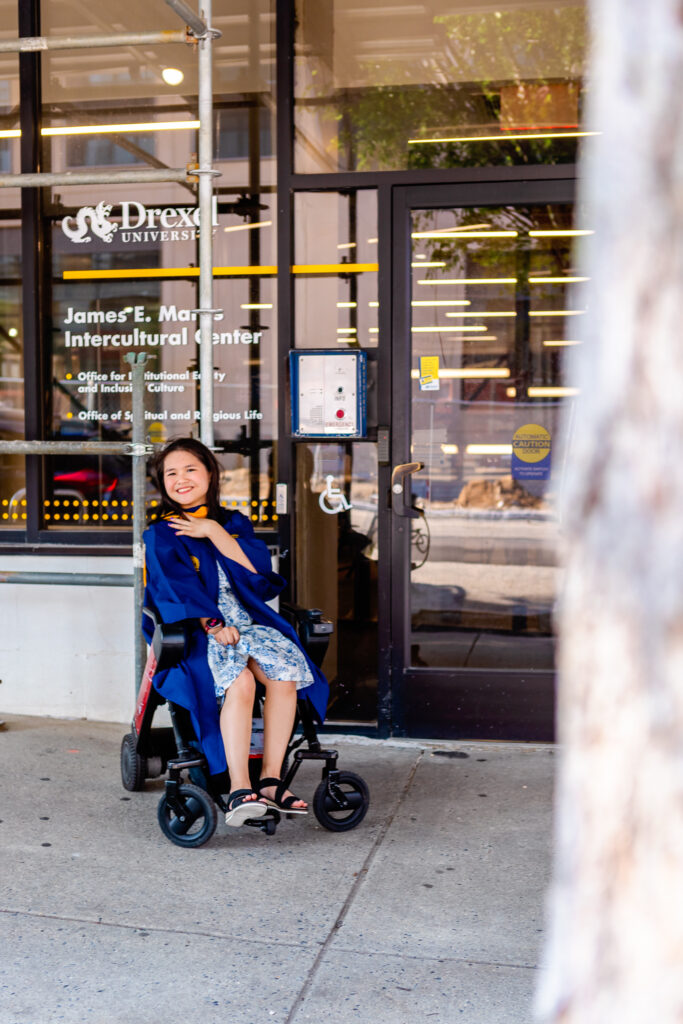 Graduate wearing academic regalia sitting in her wheelchair on campus at Drexel University, smiling proudly