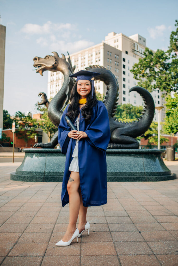 Smiling college graduate in cap and gown holding a diploma in front of the Drexel University dragon statue during a graduation photoshoot