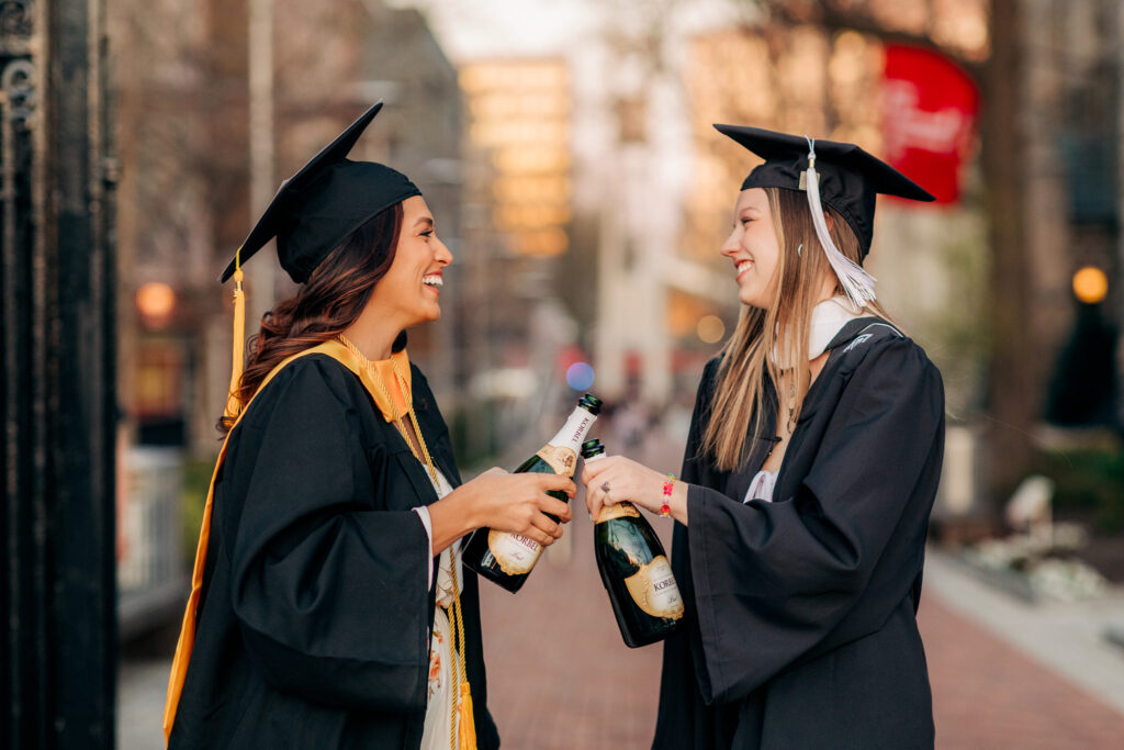 Two graduates clink champagne bottles while celebrating together on campus.